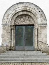 Blick auf einen Kircheneingang mit Steinstufen, Metalltüren und gemauerter, grünlich grauer Umrandung mit aufgesetztem Reliefbogen als Abschluss.
