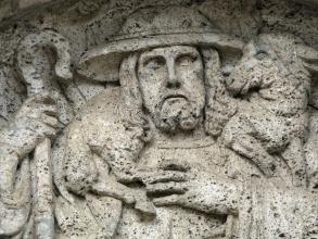 Detailaufnahme eines aus grünlich grauem Gestein gefertigten Reliefs. Das Relief stellt einen Schäfer mit Hirtenstab dar, der ein Schaf auf der Schulter trägt.