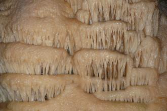 Blick in das Innere einer Tropfsteinhöhle, mit gelblich grauen Tropfsteingebilden, die - ähnlich einem Wasserfall - in mehreren Stufen nach unten wachsen. Auch eine Ähnlichkeit mit Zahnreihen von Fabelwesen ist vorhanden.
