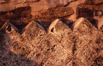 Detailaufnahme einer rötlich braunen Steinmauer mit davor angebrachter, teilweiser Verblendung durch würfelförmige Bausteine in gleicher Farbe. Die Verblendung verläuft im Zickzack-Muster; dahinter ist noch die ursprüngliche Mauer sichtbar.