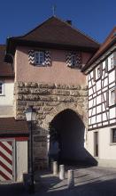 Blick auf ein mittelalterliches Stadttor, ausgeführt als viereckiger Turm mit gemauerter Durchfahrt. Das Tor hat kleine Fenster mit bemalten Läden, ein rotes Dach und einen Fachwerkanbau auf der rechten Bildseite.