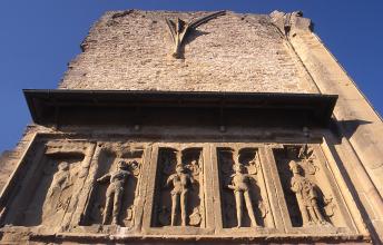 Fragment einer Hausmauer aus rötlichem bis gelblichem Gestein. Im unteren Teil verläuft eine Reihe von fünf Reliefbildern mit Figuren. Darüber ist ein schmales Vordach angebracht.