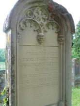 Das Foto zeigt einen grünlich grauen Grabstein mit gerundetem, verziertem Kopfteil und hebräischer Inschrift.