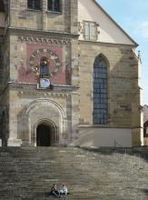 Teilansicht eines Kirchturmes aus hellem Mauerwerk mit romanischem Eingang und farbig hinterlegter Uhr (links), dem Seitenschiff mit hohem, gotischem Fenster (rechts daneben) sowie hinaufführender Freitreppe (davor).