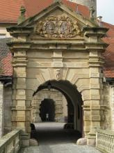 Blick auf das aus gelblich grauem Stein gefertigte äußere Tor eines Schlossbaues, mit tunnelartigem Durchgang, Giebelaufbau, Figuren und Wappentafel. Im Hintergrund ist ein weiterer Durchgang erkennbar.