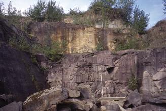 Ansicht zweier übereinanderliegender Steinbruchlager: rötliches, zerfurchtes Gestein (unten) sowie gelbliches, von Buschwerk bewachsenes Gestein darüber.