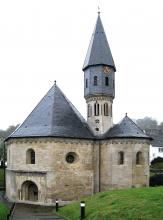 Blick auf eine dreiteilige Kirche mit gelblich grauem Mauerwerk und blaugrauen, spitzen Dächern. Die Kirche besteht aus zwei burgähnlichen Turmbauten rechts und links mit Eingang und Treppe links sowie einem mittig aufsteigenden, höheren Turm.