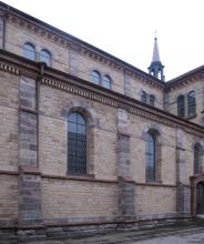 Seitenschiff einer Kirche aus gelblich grauem Mauerwerk. Fensterbögen, Stützpfeiler, Sockel und Simse sind teils bläulich, teils rötlich abgesetzt.