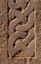 Nahaufnahme ockerbraunen, feinkörnigen Gesteins, aus dem ein von Rändern eingefasstes Muster herausgearbeitet ist. Das Muster erinnert an ineinander verschlungene Bänder.