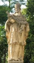 Das Foto zeigt eine größere, aus gelblich grauem Gestein gefertigte Heiligenfigur. Die Figur hält ein Kreuz mit Christus in den Händen. Im Hintergrund stehen hohe Bäume.