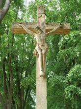Nahaufnahme eines Friedhofskreuzes mit Christusfigur aus gelblich grauem Stein. Im Hintergrund stehen hohe, dicht belaubte Bäume.