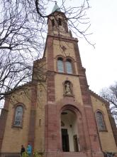 Blick von unten nach oben auf eine Kirche mit Eingangsportal, Turm und seitlichen Anbauten. Die Kirche ist aus gelblichem Mauerwerk gebaut, mit rötlichen Einfassungen an Ecken, Portal und Fenstern.