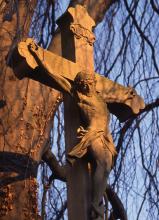 Nahaufnahme eines Kreuzes mit Christusfigur, aus (durch den Schein der tiefstehenden Sonne) rötlichem Gestein. Im Hintergrund ein Baumstamm (links) und tiefhängende kahle Äste und Zweige.