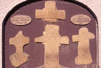 Nahaufnahme von vier Steinkreuzen, rötlich bis gelblich, vor violettem Untergrund. Die Kreuze wurden zum Gedächtnis an die Ebringer Kirchweihe im Jahre 1495 gefertigt. 