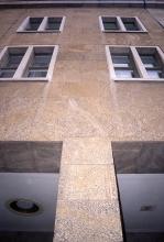 Aufwärts gerichteter Blick auf eine Gebäudefassade aus gelblich grauen Steinplatten. Unten teilt eine Säule die Fassade, darüber verlaufen Fensterreihen.