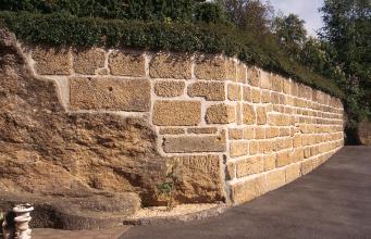Blick auf eine zum Hintergrund hin abknickende steinerne Gartenmauer mit bewachsener Krone. Links verbindet sich die Mauer mit den Ausläufern eines alten Steinbruches.