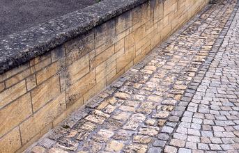 Blick auf eine gelbliche Steinmauer (links im Bild) sowie einen rechts davor verlaufenden, gepflasterten Rinnstein mit leicht abgerundeten Steinen.