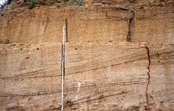 Teilansicht einer Steinbruchwand mit getrepptem Aufbau. Durch das gelblich graue Gestein verlaufen schräge, leicht wellige Streifen. Rechts ist zudem ein dünner Riss erkennbar. Links der Bildmitte ist ein Maßstab angelehnt.