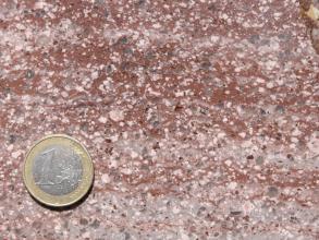 Detailaufnahme eines Gesteins mit unterschiedlichen Farben. Oben zeigen sich rötlich braune Flächen, unten rosa und graue Streifen. Auch zahlreiche Einsprenglinge sind erkennbar. Am linken, unteren Bildabschnitt liegt eine Eu­ro­mün­ze als Maßstab.
