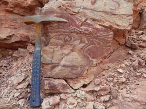 Das Bild zeigt im Wechsel rotbraune bis graugrünliche Schlieren im Gestein. Im linken Bereich des Bildes ist ein Geologenhammer als Maßstab platziert.