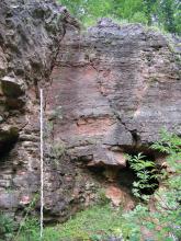 Blick auf eine über Eck gehende Steinbruchwand, bläulich bis rötlich, mit einer höhlenartigen Öffnung rechts unten. Links lehnt eine Messlatte am Gestein.