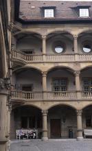 Blick auf die dreistöckige Fassade eines Gebäudes aus hellbraunem Mauerwerk mit Säulen in jedem Stockwerk sowie Galerien.