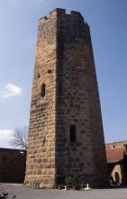 Die Aufnahme zeigt einen hohen, achteckigen Burgturm aus gelblich grauen Mauersteinen. Der Turm ist mit Zinnen bekrönt und weist zwei dunkle Fensteröffnungen auf.