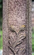 Teilansicht eines rötlich grauen, steinernen Grabkreuzes mit herausgearbeiteter Verzierung in Form einer Blume mit zwei Kelchen und Blättern. Der Stein weist zudem zahlreiche dunklere Flecken auf.