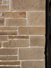 Teilansicht einer steinernen Hausmauer mit Abschluss rechts. Das Gestein ist rau und streifig, hellbraun bis rötlich braun und weist an der Ecke rechts größere Blöcke auf als links.