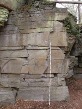 Teilansicht einer hellbraunen Steinbruchwand mit grauem Sockel sowie grünlichen Verfärbungen im oberen Bereich. Der Sockel des Gesteins zeigt zudem Einkerbungen.