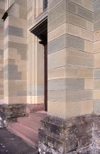 Blick auf das Eingangsportal einer Kirche mit beidseitig vorstehenden Stützpfeilern. Das Mauerwerk ist mehrfarbig, teils grünlich bis bläulich, teils hellbraun.