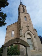 Aufwärts fotografierter Kirchturm aus hellem Mauerwerk mit Eingangsportal rechts und seitlichem Anbau links. Im Vordergrund ein steinerner Wappenbogen.