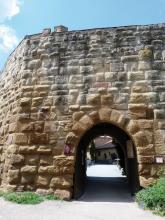 Blick auf eine Burgmauer aus wulstigen, hellbraunen bis schwarzgrauen Mauersteinen. Die Mauer hat rechts einen tunnelartigen Durchgang; dahinter sind weitere Bauten erkennbar.