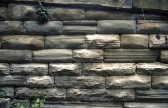 Teilansicht einer Steinmauer mit dunkelgrauen bis graubraunen Mauersteinen, zum Teil mit weißlichen Verfärbungen.