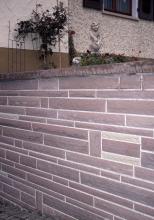 Teilansicht einer rötlich grauen Steinmauer, die quer zu Hauswänden steht.
