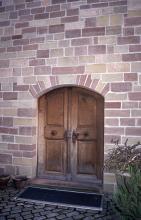 Blick auf eine Gebäudewand aus mehrfarbigen Mauersteinen. Eine Doppeltür aus Holz bildet den Eingang.