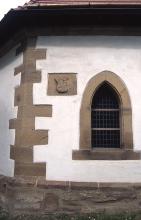 Teilansicht eines kirchlichen Gebäudes mit Ecksteinen, Fensterumrandung und Wappen aus hellbraunem Mauerwerk. Sockel dunkelgrau, Wand weiß verputzt.