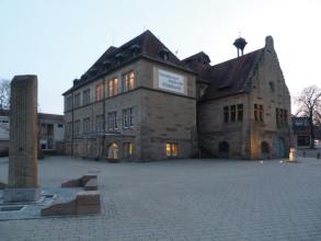 Blick auf zwei versetzt nebeneinander stehende Gebäude aus graubraunem Mauerwerk. Das Gebäude links trägt ein Schild mit der Aufschrift „Steinhauer Museum Mühlbach“. Es ist Abend, und ein paar Fenster sind erleuchtet. Links vorne steht eine Steinsäule.