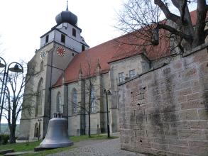 Blick auf Turm und Hauptschiff einer Kirche aus hellbraunem Mauerwerk. Der Turm hat einen weiß verputzten Aufsatz sowie ein Zwiebeldach. Im Vordergrund rechts eine Steinmauer, links ist eine große Glocke ausgestellt.