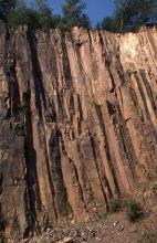 Blick auf eine hohe, rötlich braune Steinbruchwand mit leicht schräg verlaufender Säulenstruktur. 