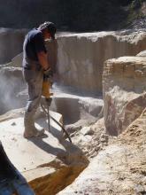 Blick auf einen Arbeiter in einem Steinbruch, der – auf einem Steinblock stehend – diesen mit einem Bohrgerät bearbeitet. Im Hintergrund Steinbruchwände.