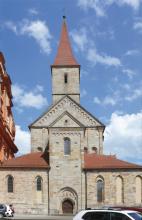 Blick auf eine Kirche mit Eingang, Hauptbau, seitlichen Anbauten und Turm. Die Kirche besteht aus hellgrauem bis hellbraunem Mauerwerk. Die Dächer tragen rote Ziegel.