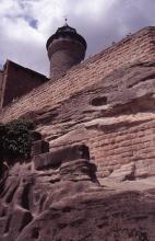 Aufwärts gerichteter Blick auf eine rötlich braune, waagrecht abgeteilte Burgmauer sowie einen darüber aufragenden runden Turm. Die Mauer unterbrechen rohe Gesteinsbänke, die gefurcht und gefaltet sind.