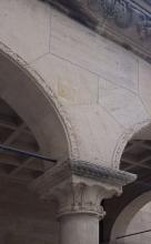 Blick auf einen aus mehreren Teilen bestehenden Säulenaufsatz, rötlich grau mit Verzierungen am Rand und geformt wie ein steinerner Brückenbogen.