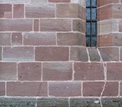 Teilansicht einer Kirchenfassade aus rötlich grauem Mauerwerk über grünlich grauem Sockel. Rechts oben ist in einer Fensterbucht ein Bleiglasfenster eingelassen.