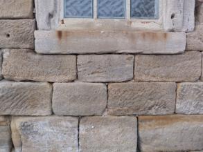 Teilansicht einer gemauerten Hausfassade aus rötlich grauen Steinen mit Anschnitt eines Fensters am oberen Bildrand. Die Mauersteine sind gefurcht, an den Kanten gerundet und gefleckt. Auch die steinerne, hellere Fensterbank weist Verfärbungen auf.