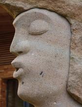 Nahaufnahme vom Kopfteil einer stilisierten Steinfigur. Das Gestein des Gesichtes ist rötlich grau und streifig, mit dunkleren Einschlüssen an Kinn und Wange. Das Haar - ebenfalls aus Stein - ist etwas bräunlicher.