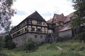 Blick auf mehrere ältere, miteinander verbundene Fachwerkgebäude. Der Unterbau der Häuser besteht aus grauem Mauerwerk. Die Gebäude sind von Bäumen, Büschen und einer Wiese umgeben.