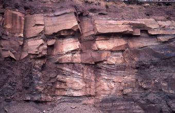 Blick auf eine Gesteinswand mit rötlichen bis violetten Gesteinen. Unterhalb der Bildmitte weisen die Steine eine streifige Färbung auf.