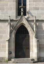 Teilansicht einer Kirche mit schmalem, aus hellem Gestein bestehendem und von Stützpfeilern, kleinen Türmchen sowie einem Spitzdach umrahmtem Eingangsportal.